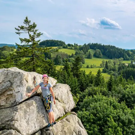 Žena s horolezeckou výstrojí a helmou stojí na skalnatém výběžku a usmívá se, v pozadí je zelený les a kopce pod částečně zataženou oblohou.