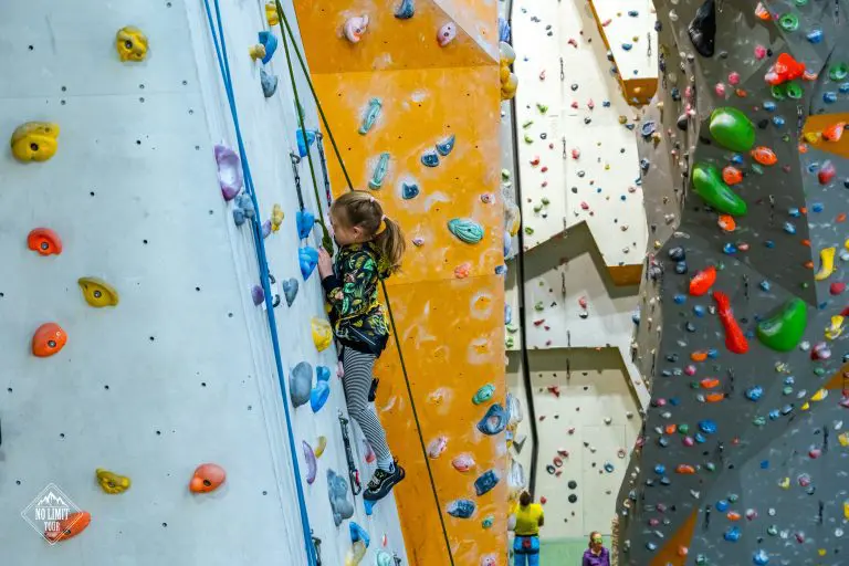 Mladá dívka v pestrobarevném tričku a pruhovaných kalhotách leze po pestrobarevné vnitřní horolezecké stěně, jištěná postrojem a lanem. Na pozadí jsou vidět různé lezecké cesty a chyty na stěnách.