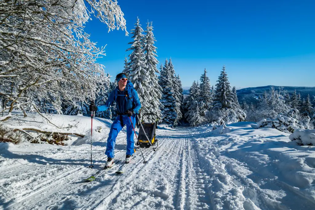 Osoba v modrém zimním oblečení na běžkách na zasněžené trase, táhnoucí dětský přívěs přes sluncem ozářený les se zasněženými stromy pod jasně modrou oblohou.