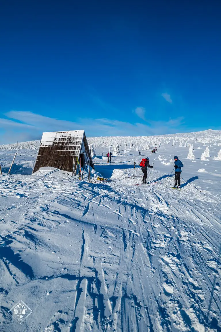 Lyžaři v zimní výstroji stojí u malé dřevěné chaty v zasněžené krajině pod jasnou modrou oblohou a v pozadí jsou zasněžené stromy.