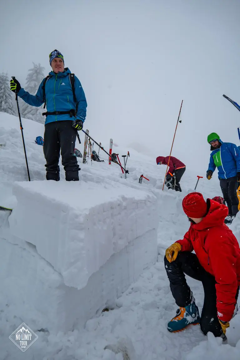 Skupina lidí v zimní výstroji stojí a klečí kolem velkého obdélníkového bloku sněhu během zasněženého mlhavého dne, v pozadí jsou vidět lyžařské hůlky a vybavení.