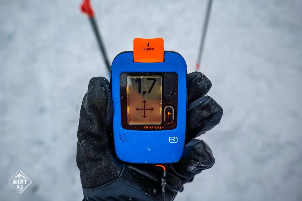 Ruka v rukavici drží modrý lavinový maják, na jehož displeji se zobrazuje hodnota 1,7. V pozadí je vidět sníh a lyžařské hůlky.