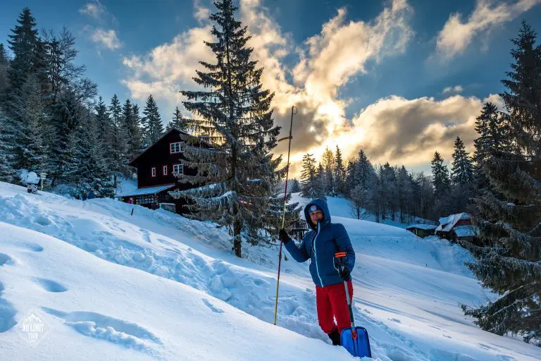Osoba v zimním oblečení stojí v hlubokém sněhu s lyžařskou holí a modrou lopatou, obklopená zasněženými stromy a dřevěnými chatkami pod částečně zataženou oblohou při západu slunce.