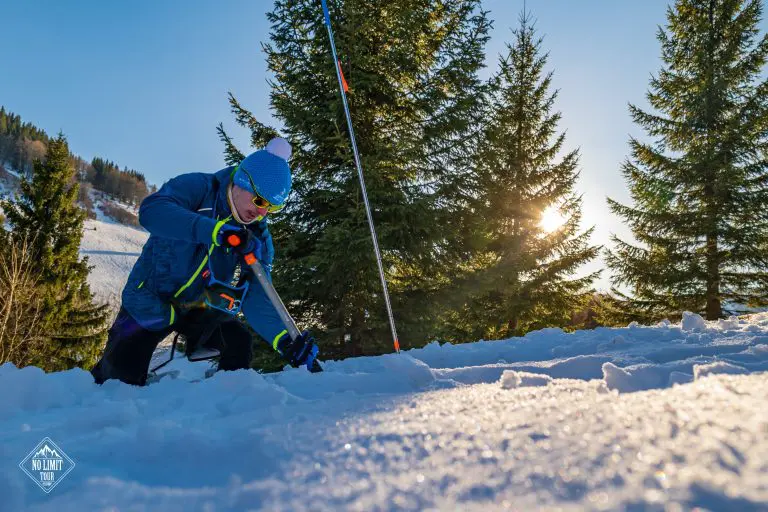 Osoba v zimní výstroji hledá sondou ve sněhu na slunném zasněženém svahu obklopeném stálezelenými stromy, mezi jejichž větvemi proniká sluneční světlo.