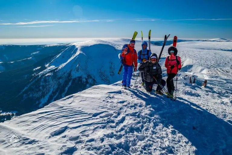 Čtyři lyžaři v zimní výstroji pózují na zasněženém horském vrcholu pod jasnou modrou oblohou, v pozadí se rozprostírají zasněžené hory a údolí.