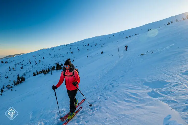 Osoba v červené bundě lyžuje při západu slunce na zasněženém horském svahu a drží lyžařské hůlky. Další osoba je vidět dále po trase. Zasněženou krajinu pod jasnou modrou oblohou lemují malé stromy.