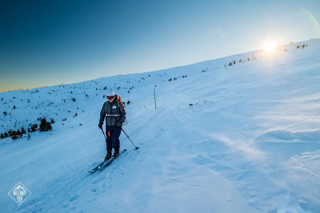 Osoba v zimní výstroji lyžuje na zasněženém svahu do kopce. Slunce je nízko nad obzorem a vrhá teplou záři na zasněženou krajinu s roztroušenými stromy.
