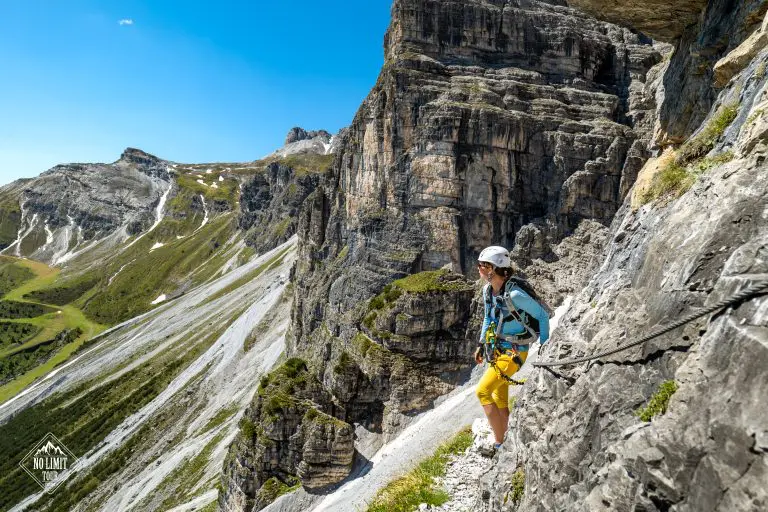 Horolezec v přilbě a sedacím úvazku stojí na úzkém skalnatém horském výstupku, drží se jistícího lana a pod jasně modrou oblohou jsou vidět strmé skály a zelená údolí.