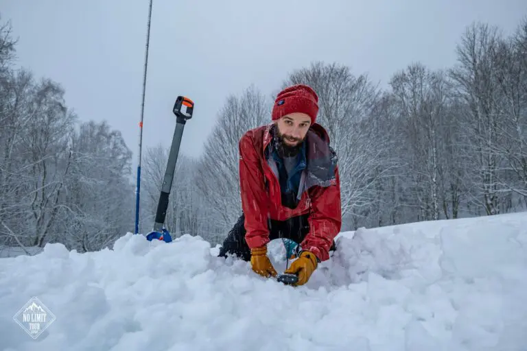 Muž v červené bundě a pletené čepici klečí v hlubokém sněhu a rukama hrabe. V pozadí jsou vidět zasněžené stromy a sloup s vybavením. Obloha je zatažená.