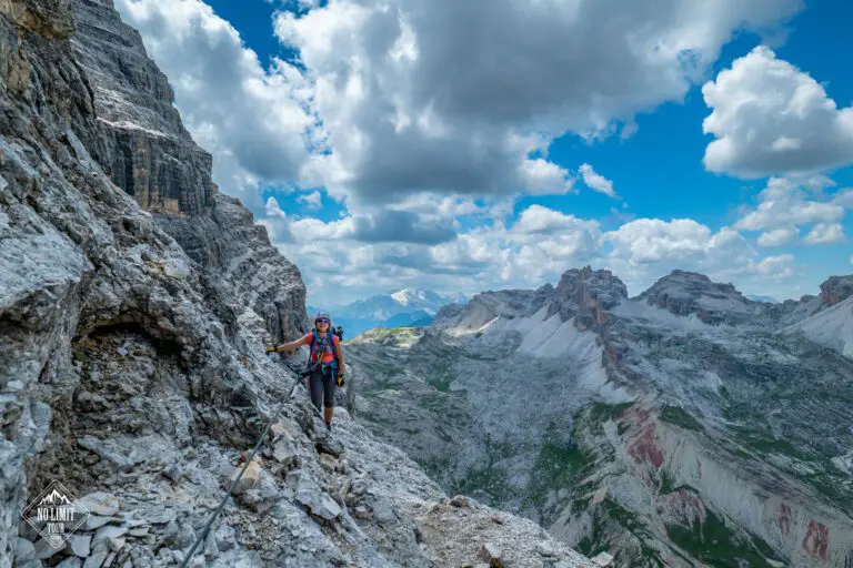 Turista v horolezecké výstroji překonává skalnatou horskou stezku pomocí jistícího lana, obklopen drsnými vrcholy a dramatickými mraky pod jasně modrou oblohou.