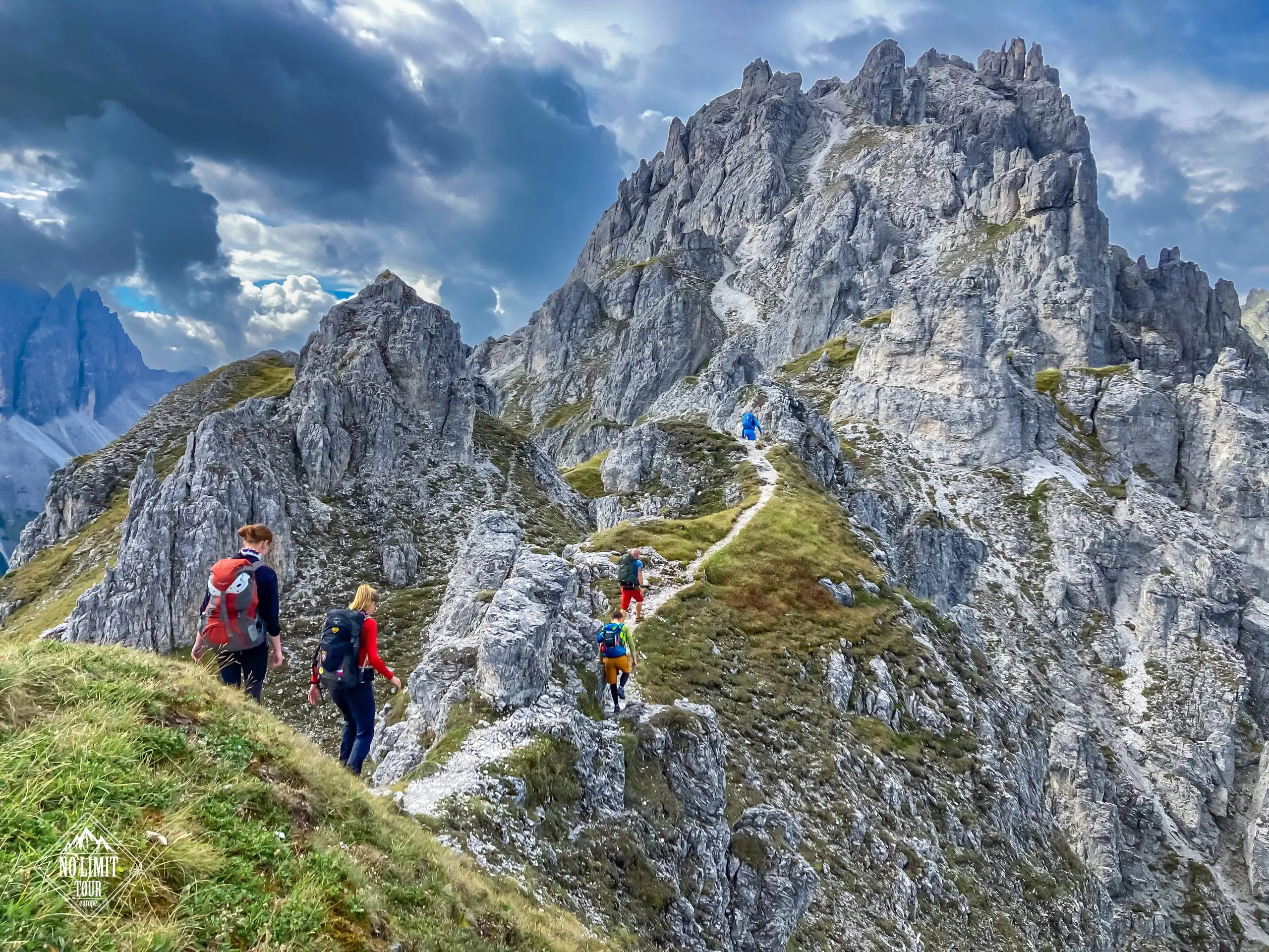 Turisté s batohy na zádech kráčejí po úzké klikaté horské stezce obklopené drsnými skalnatými vrcholy pod dramatickou zataženou oblohou.