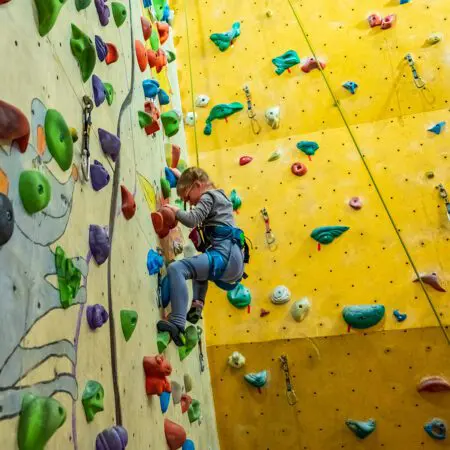 Dítě v sedacím úvazku a s horolezeckou výstrojí stoupá na vnitřní horolezeckou stěnu s barevnými chyty, soustředěně a pevně se drží. V pozadí jsou vidět další lezecké chyty a lana připevněná ke stěně.
