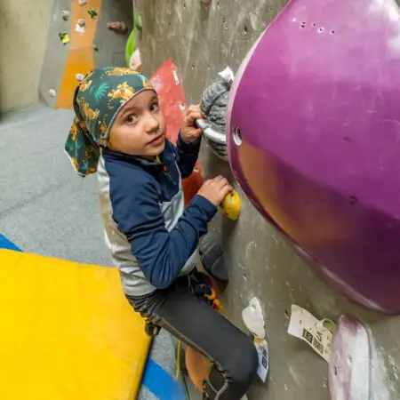 Malé dítě ve vzorovaném šátku a sportovním oblečení leze na vnitřní boulderingové stěně, chytá se barevných chytů a dívá se do kamery. Dole je vidět žlutá polstrovaná podložka.