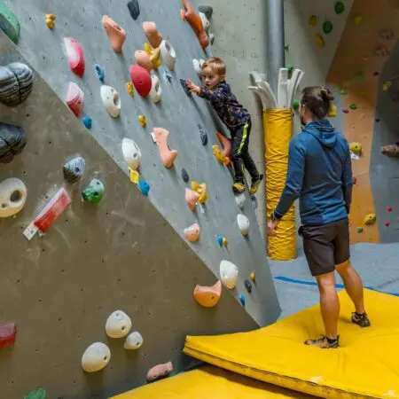 Malé dítě leze na barevnou vnitřní boulderingovou stěnu, zatímco dospělý stojí opodál na žluté podložce a je připraven mu v případě potřeby pomoci nebo ho chytit. Lezecká stěna v tělocvičně je vybavena různými chyty a opěrkami.