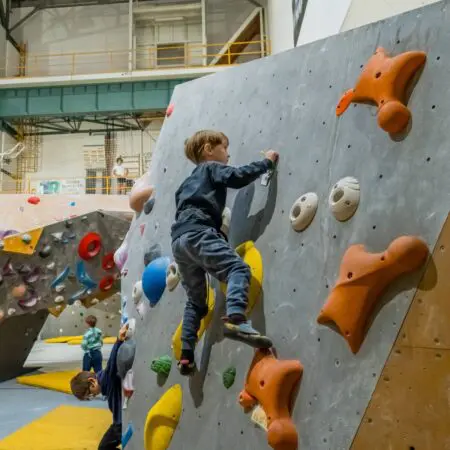 Malý chlapec leze na vnitřní boulderingové stěně s barevnými chyty, zatímco další děti lezou opodál. Prostředí je prostorná krytá lezecká tělocvična s bezpečnostními žíněnkami na podlaze.