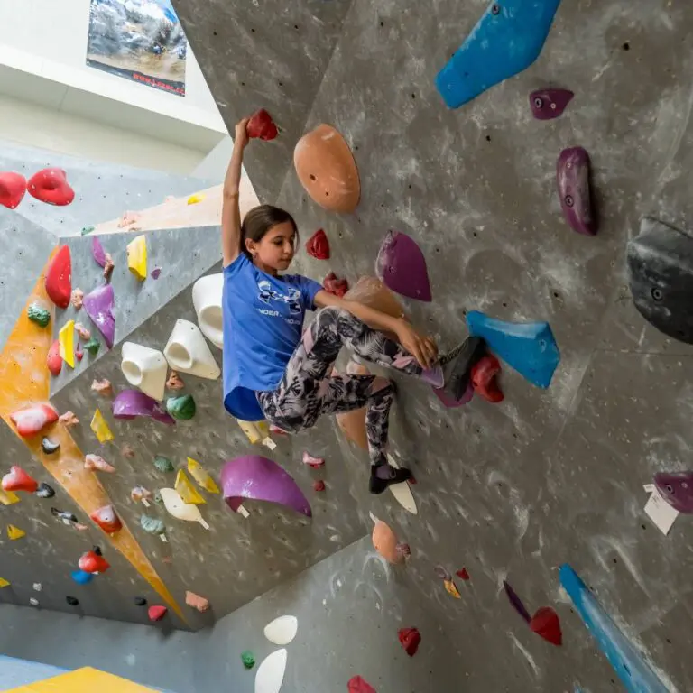 Mladá dívka v modrém tričku a vzorovaných legínách leze po vnitřní boulderingové stěně s barevnými chyty, jednou rukou se natáhne nahoru a opře se nohama o stěnu.
