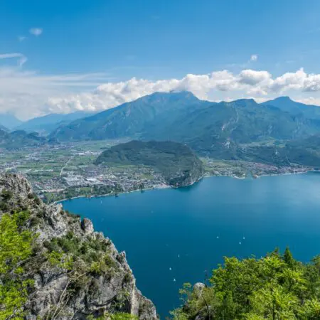 Malebný pohled na tmavě modré jezero obklopené zelenými horami a útesy, s malými loďkami na vodě a městečkem na úpatí kopců pod částečně zataženou oblohou.