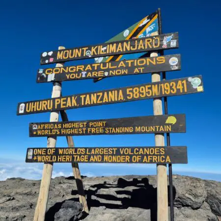 Dřevěná cedule na vrcholu Kilimandžára hlásá: Mount Kilimanjaro, Gratulujeme, Nyní jste na vrcholu Uhuru Peak, Tanzanie, 5895 m/19341 stop, a upozorňuje na jeho status nejvyššího bodu Afriky a světového dědictví.