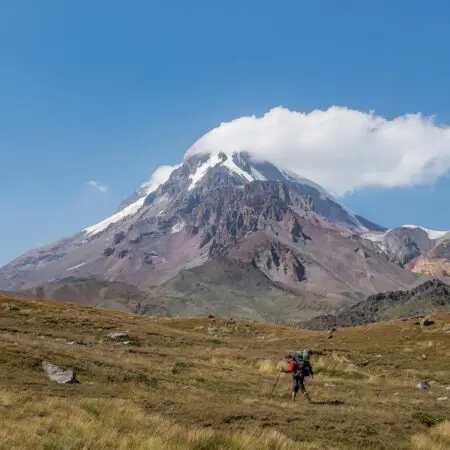 Turista s velkým batohem a trekingovými holemi kráčí travnatým terénem k vysoké zasněžené hoře pod jasně modrou oblohou, jejíž vrchol zakrývá mrak.