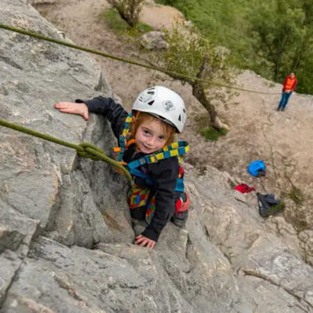 Malé dítě s helmou a úvazkem leze venku po skalní stěně, dívá se vzhůru a usmívá se. Na zemi pod ním je vidět dospělý s horolezeckým vybavením.