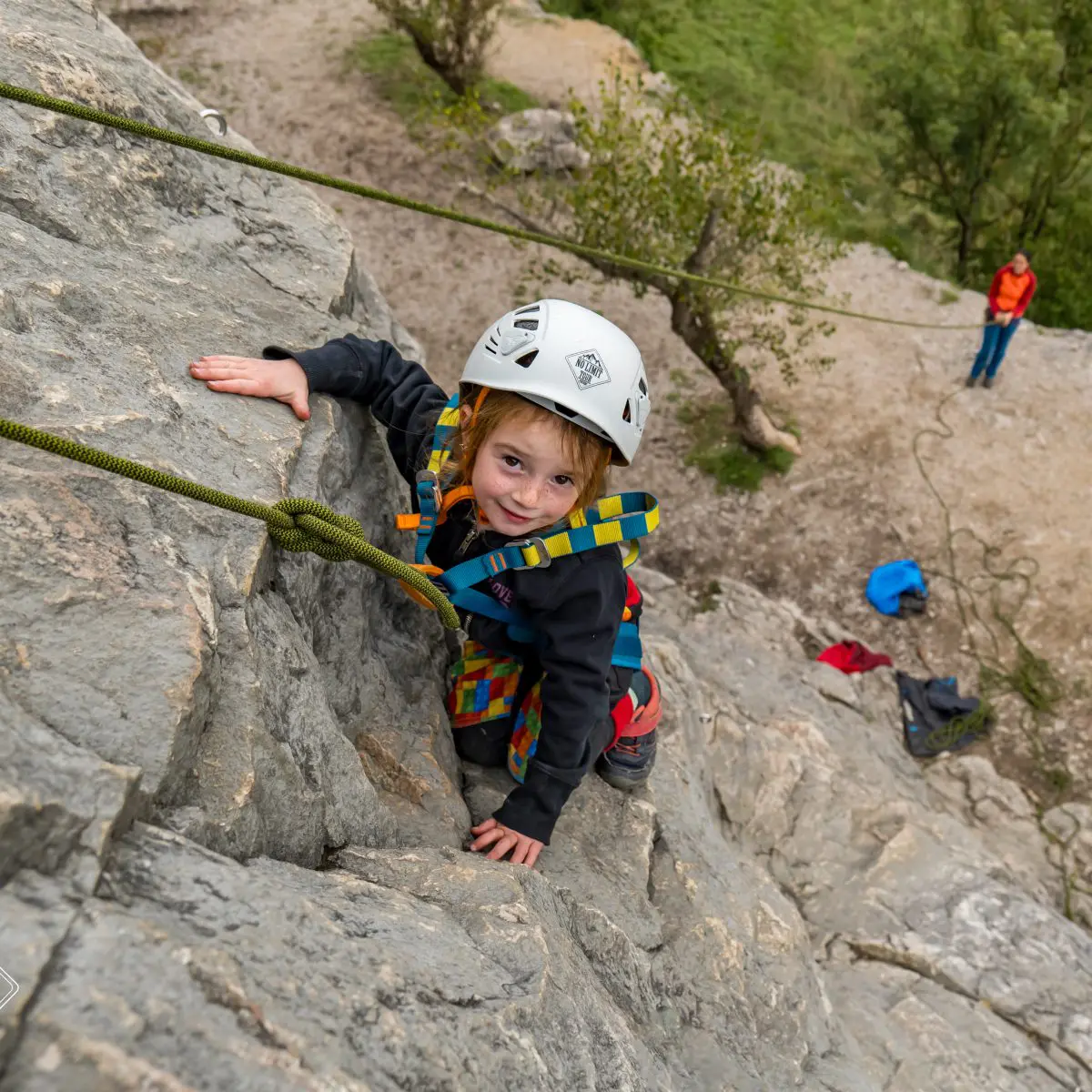 Malé dítě s helmou a úvazkem leze venku po skalní stěně, dívá se vzhůru a usmívá se. Na zemi pod ním je vidět dospělý s horolezeckým vybavením.