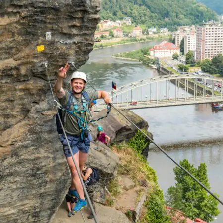 Muž v přilbě a postroji se usmívá a dělá znamení míru, zatímco leze po skalnatém útesu nad řekou, v pozadí je vidět město, most a zelené kopce. Pod ním jsou další horolezci.