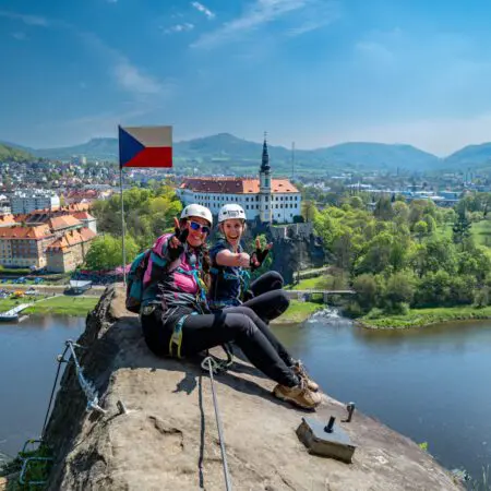 Dva usměvaví horolezci v helmách a s výstrojí sedí na skalní římse s českou vlajkou a mají výhled na řeku, budovy a zelenou krajinu pod modrou oblohou.