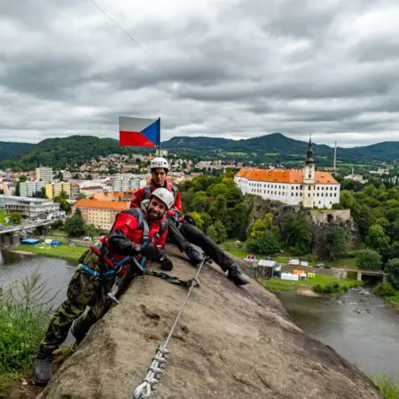 Dva lidé v helmách a s výstrojí jsou připoutáni lany na skalním útesu a usmívají se. Na jednom postroji je připevněna česká vlajka. V pozadí je pod zataženou oblohou vidět malebné město, řeka a historická budova s věží.