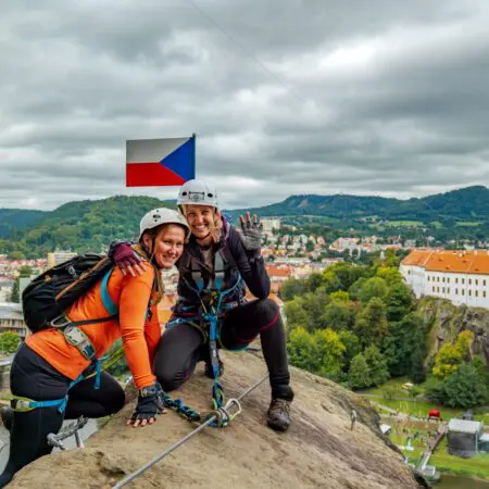 Dvě usměvavé ženy v horolezecké výstroji pózují na skalní římse s bezpečnostními lany, na hlavách mají helmy a rukavice. Za nimi je česká vlajka a v pozadí malebné město a svěže zelené kopce.