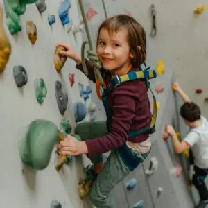 Malé dítě s úvazkem se usmívá při lezení na vnitřní skalní stěně. V pozadí leze další dítě. Stěna je vybavena barevnými úchyty a bezpečnostními lany.