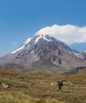 Turista s velkým batohem a trekingovými holemi kráčí travnatým terénem k vysoké zasněžené hoře pod jasně modrou oblohou, jejíž vrchol zakrývá mrak.