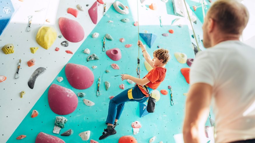 Father and teenager son at indoor climbing wall hall. Boy is hanging on the rope using a climbing harness and daddy belaying him on the floor using a belay device. Happy parenting concept image.