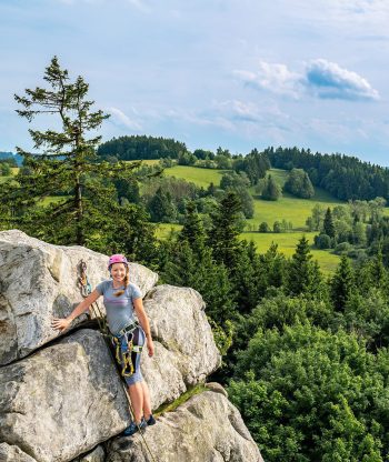 Osoba v horolezecké výstroji a přilbě stojí na velkém skalním útvaru a usmívá se do kamery, v pozadí jsou zelené kopce, lesy a částečně zatažená obloha.