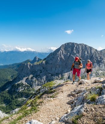 Dva turisté s batohy na zádech kráčejí za slunečného dne po skalnaté horské stezce, obklopeni rozeklanými vrcholky, zelenými lesy a jasnou modrou oblohou v pozadí.