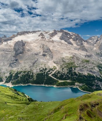 Úchvatná horská krajina se skalnatými vrcholky, sněhovými skvrnami, tyrkysovým jezerem dole, zelenými travnatými kopci v popředí a částečně zataženou oblohou nahoře.