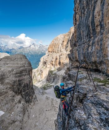 Horolezec v přilbě a sedacím úvazku stoupá po kovových příčkách na strmou skalnatou stěnu hory s dramatickými útesy a vzdálenými horami viditelnými pod jasně modrou oblohou.