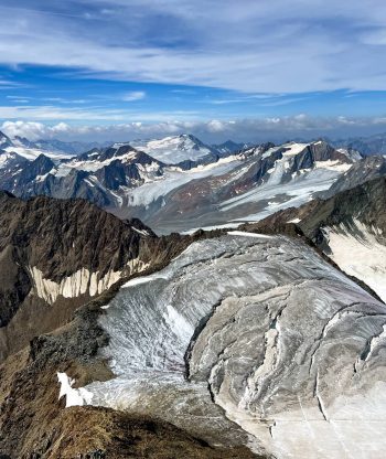 Pohled na členité zasněžené hory s ledovci a skalnatými hřebeny pod částečně zataženou modrou oblohou, která ukazuje rozlehlou alpskou krajinu.