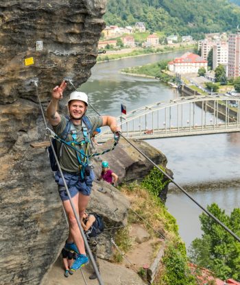Muž v přilbě a postroji se usmívá a dělá znamení míru, zatímco leze po skalnatém útesu nad řekou, v pozadí je vidět město, most a zelené kopce. Pod ním jsou další horolezci.