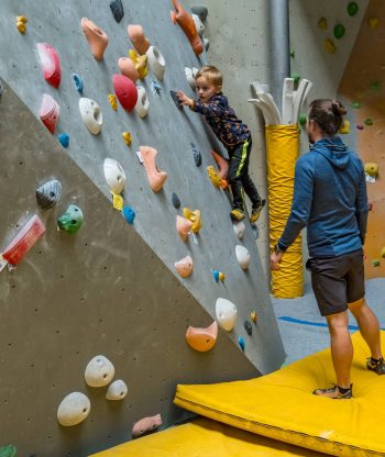 Malé dítě leze na barevnou vnitřní boulderingovou stěnu, zatímco dospělý stojí opodál na žluté podložce a je připraven mu v případě potřeby pomoci nebo ho chytit. Lezecká stěna v tělocvičně je vybavena různými chyty a opěrkami.